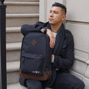 Man holding a black backpack with brown leather accents and visible branding, 'Chef Sac' sitting against a stone wall 