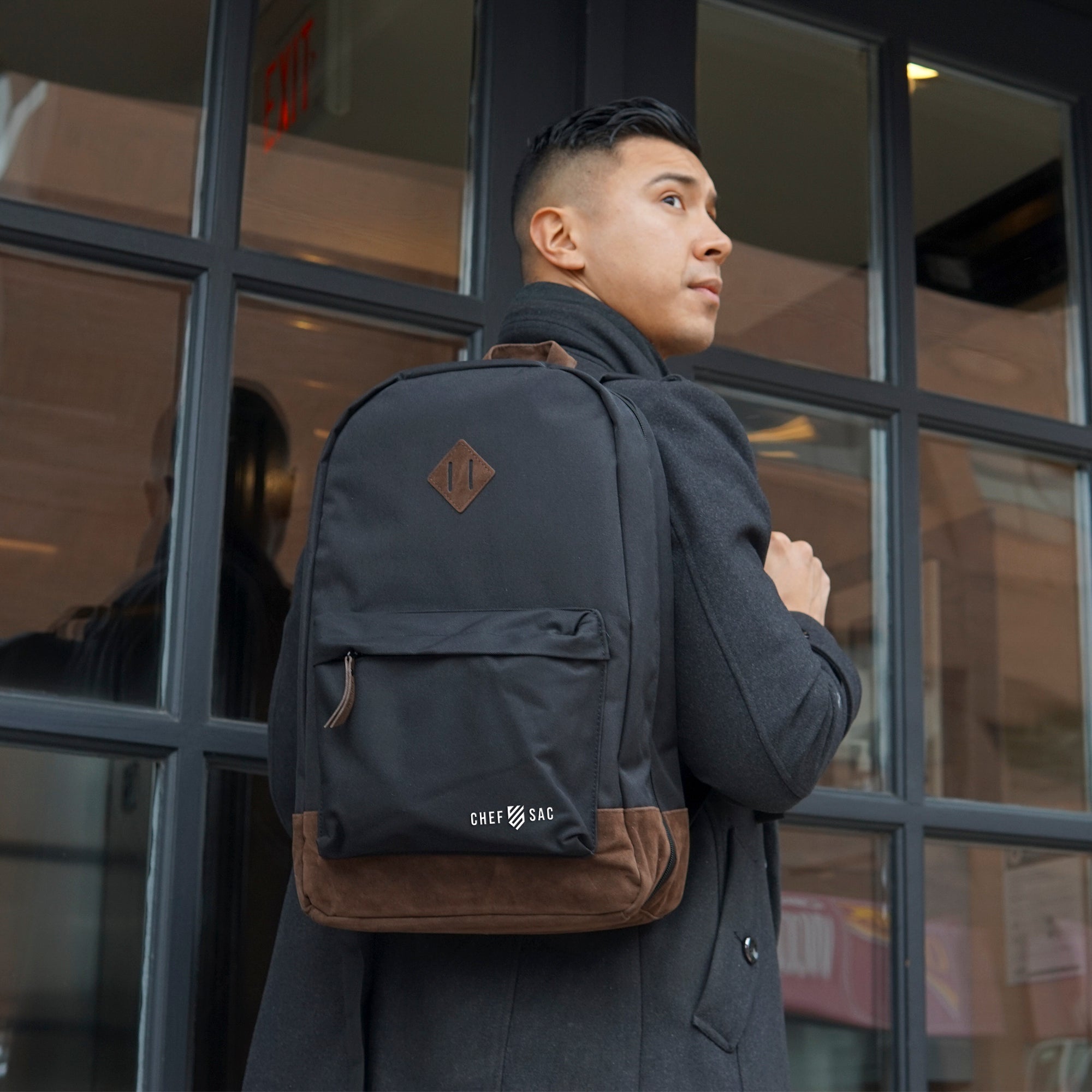 Man wearing a black backpack with brown leather accents, and visible branding, 'Chef Sac' while standing in front of a glass door.