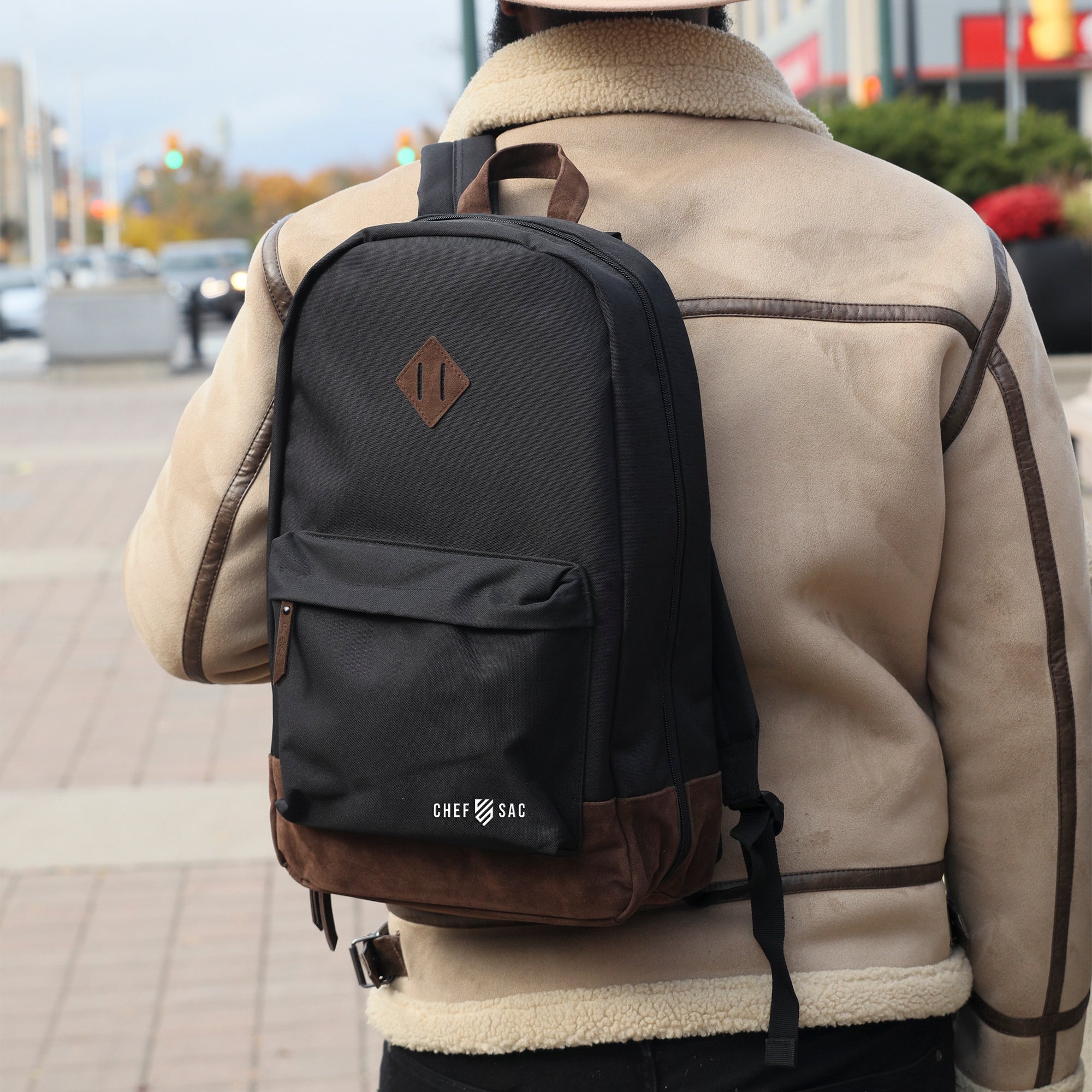 Person wearing a black backpack with brown leather accents on a city street and visible branding, 'Chef Sac'